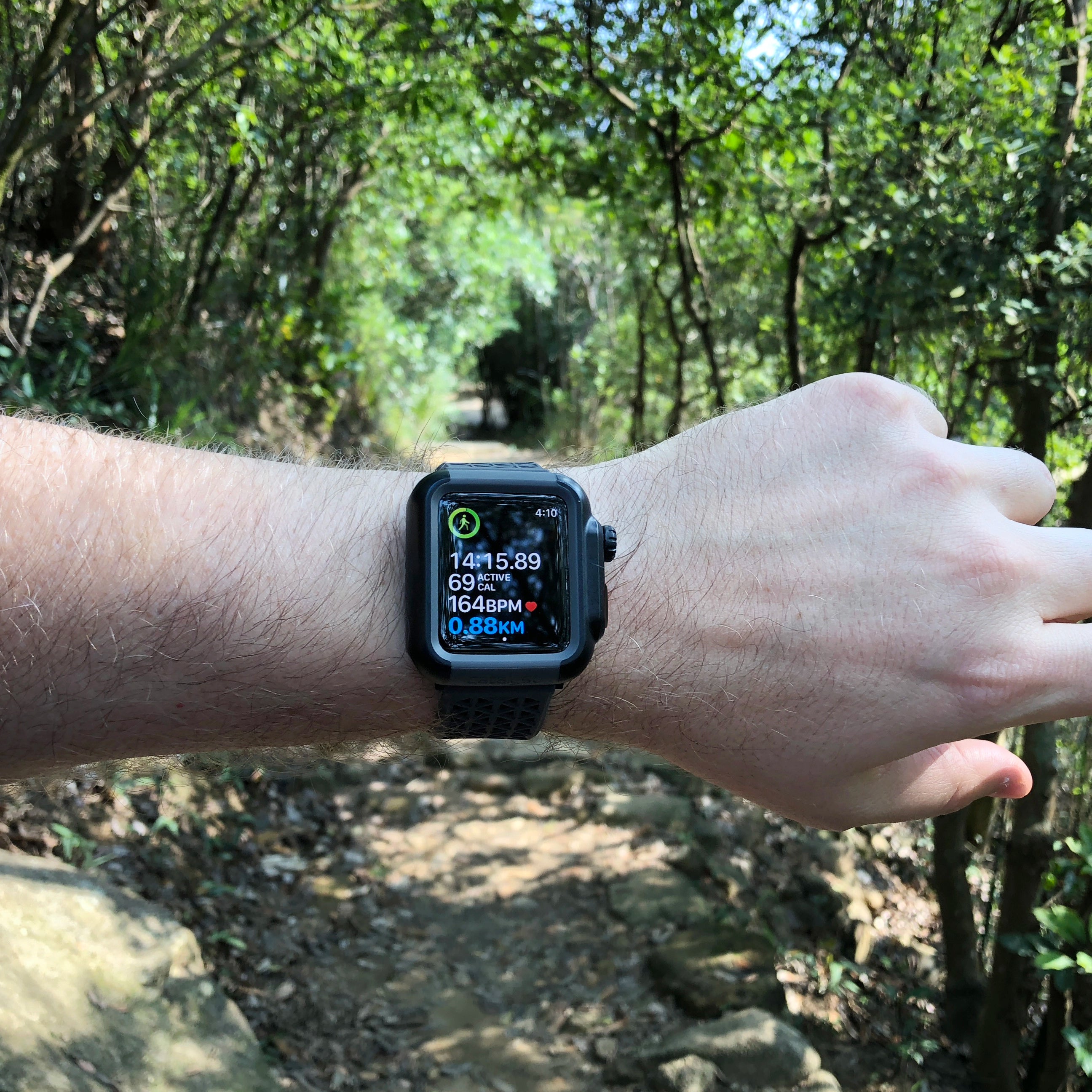 Using a Catalyst Apple Watch waterproof case in black, the left wrist displays fitness metrics like steps and heart rate while hiking on a wooded trail.