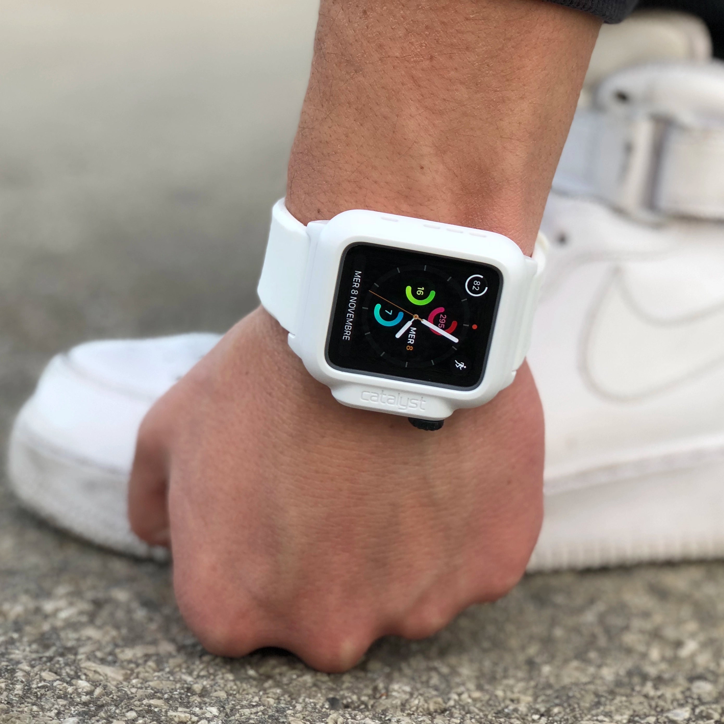 A person wearing an Apple Watch with a Catalyst white waterproof case, displaying fitness data, rests their hand on a textured surface. They're also wearing sleek white shoes.