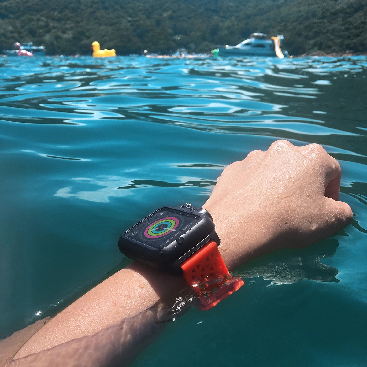 A person's arm, featuring a Catalyst Apple Watch - Waterproof Case with a red band, gracefully extends in water surrounded by inflatables and distant trees.
