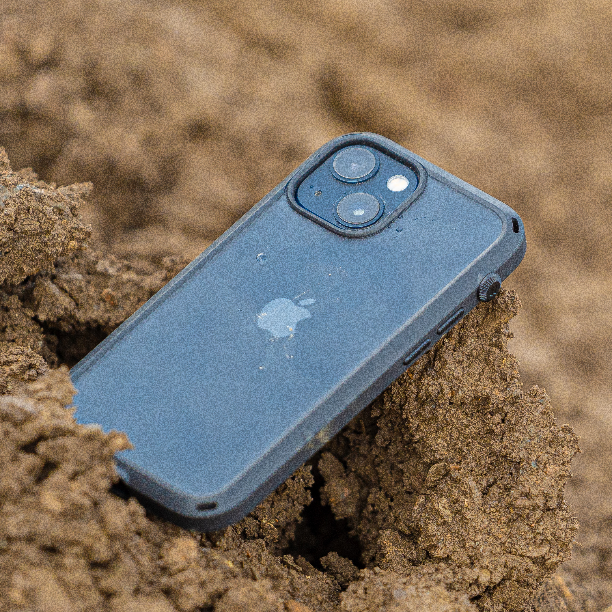An iPhone with a Catalyst Drop Proof Case in black, featuring dual cameras, is partially buried in soil.