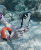 A close up of a woman taking a photo underwater in a black Catalyst waterproof case. The iPhone is secured to her wrist through a Floating Lanyard in orange.
