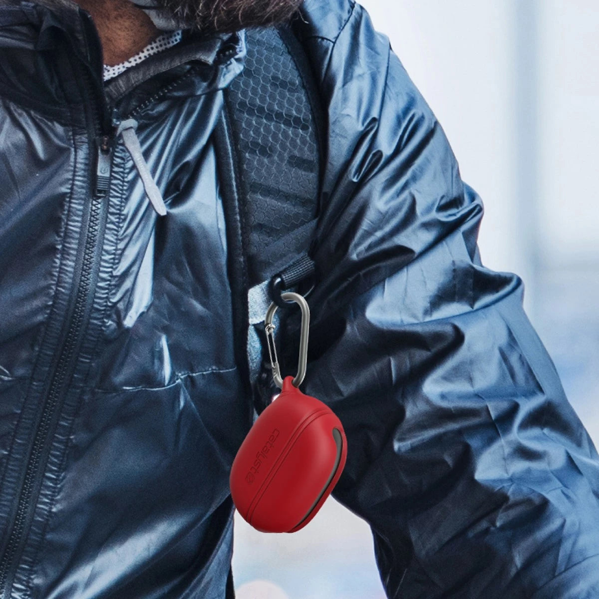 A person in a shiny blue jacket sports a red carabiner, a small red pouch on their backpack strap, and a Beats Studio Buds Waterproof Case by Catalyst that seals out water and dirt.