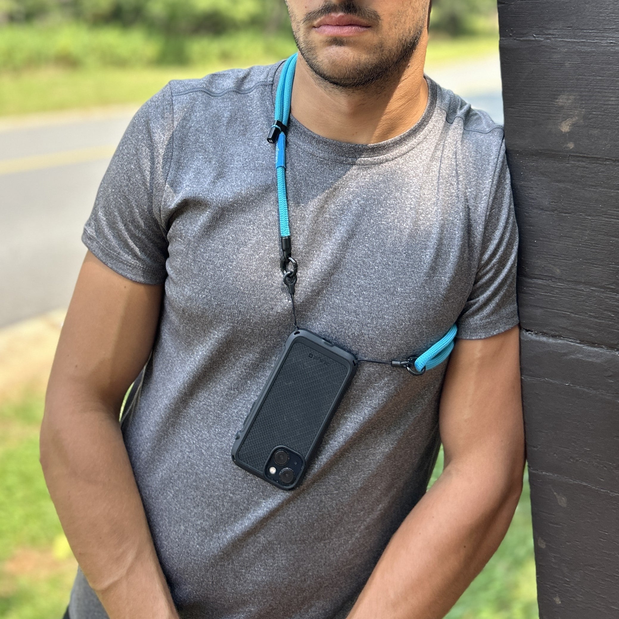A man leans against a wooden post outdoors, wearing a gray shirt. His black Catalyst iPhone Vibe Case, featuring drop protection, hangs from a blue lanyard around his neck.