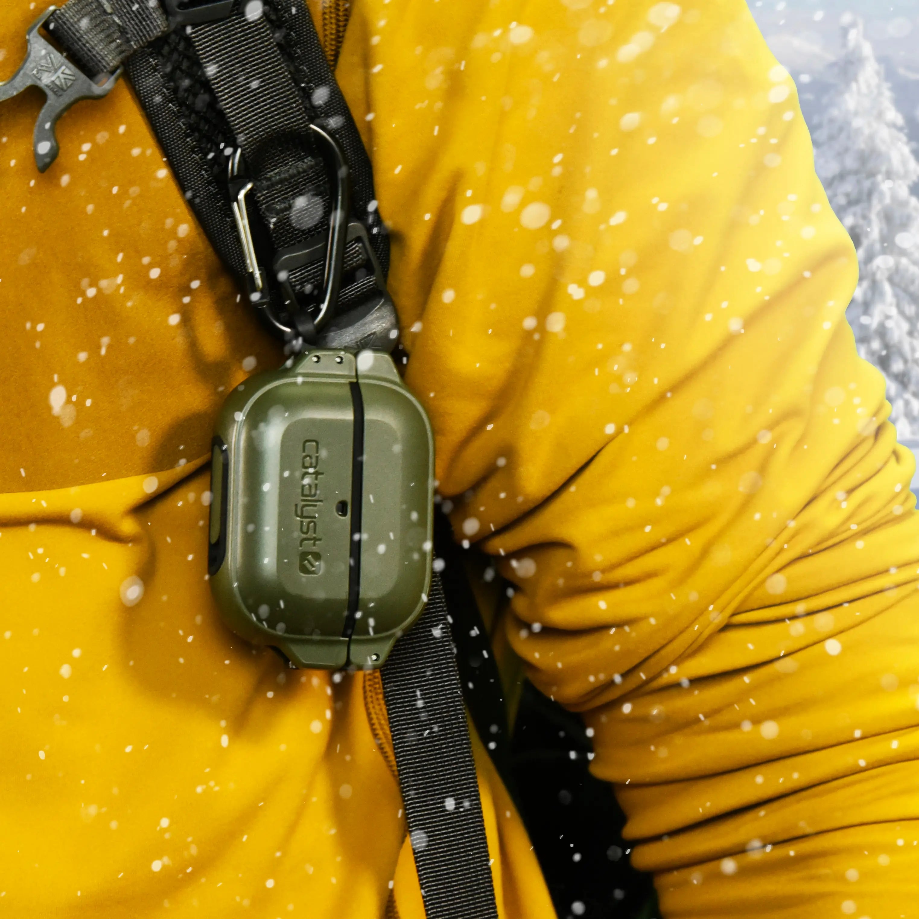 Wearing a yellow jacket, a person has an Army Green Catalyst AirPods Pro Waterproof Case with total protection against water and impacts attached to a black strap. Snowflakes gently fall around, showcasing its ability to guard against dirt, dust, germs, and drops.