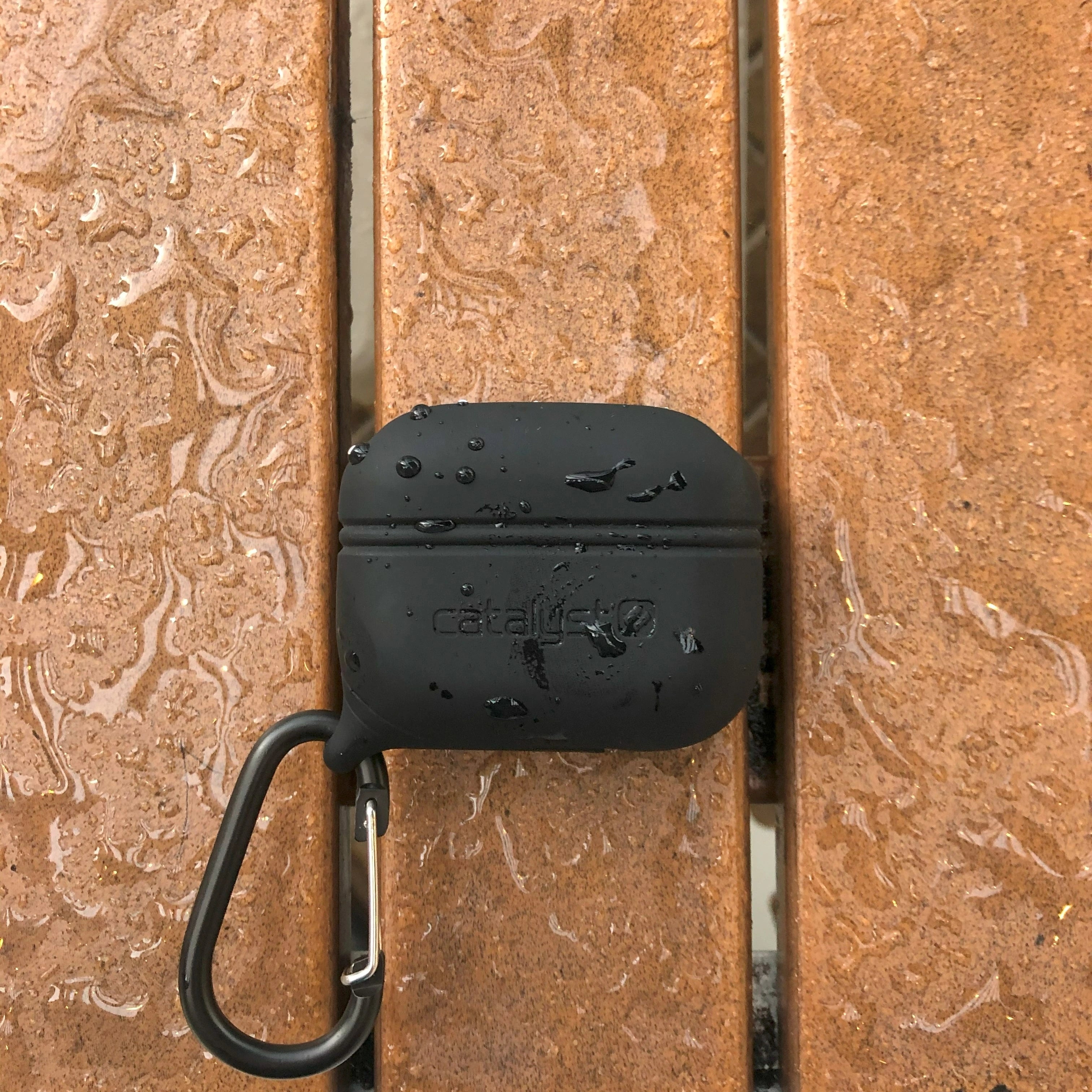 A Catalyst AirPods Pro (Gen 2/1) black waterproof case with a carabiner sits elegantly, covered in water droplets, on a wet wooden surface.