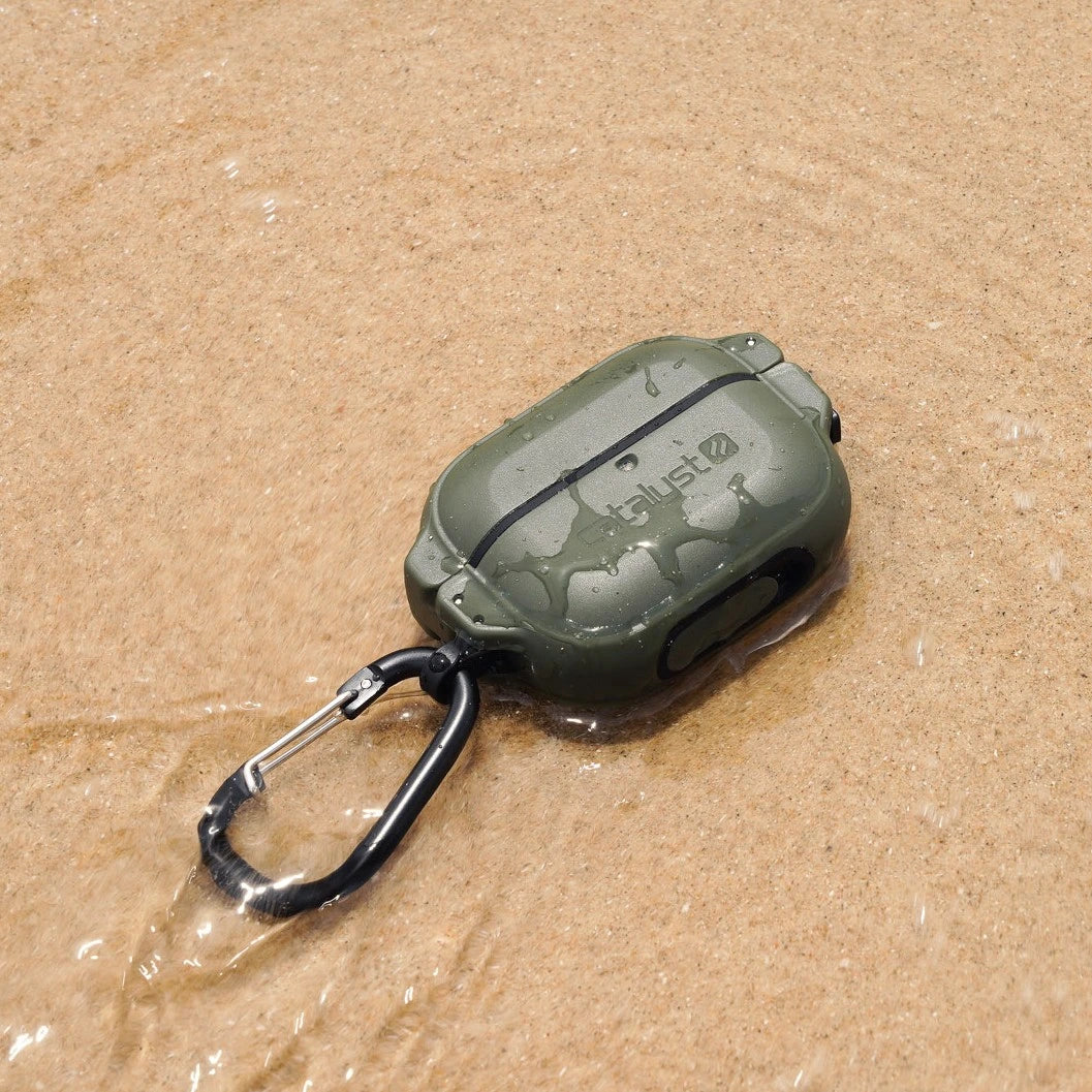 Catalyst's Army Green AirPods Pro (Gen 2/1) - 100m Waterproof Case with carabiner rests on wet sand, gently surrounded by water.