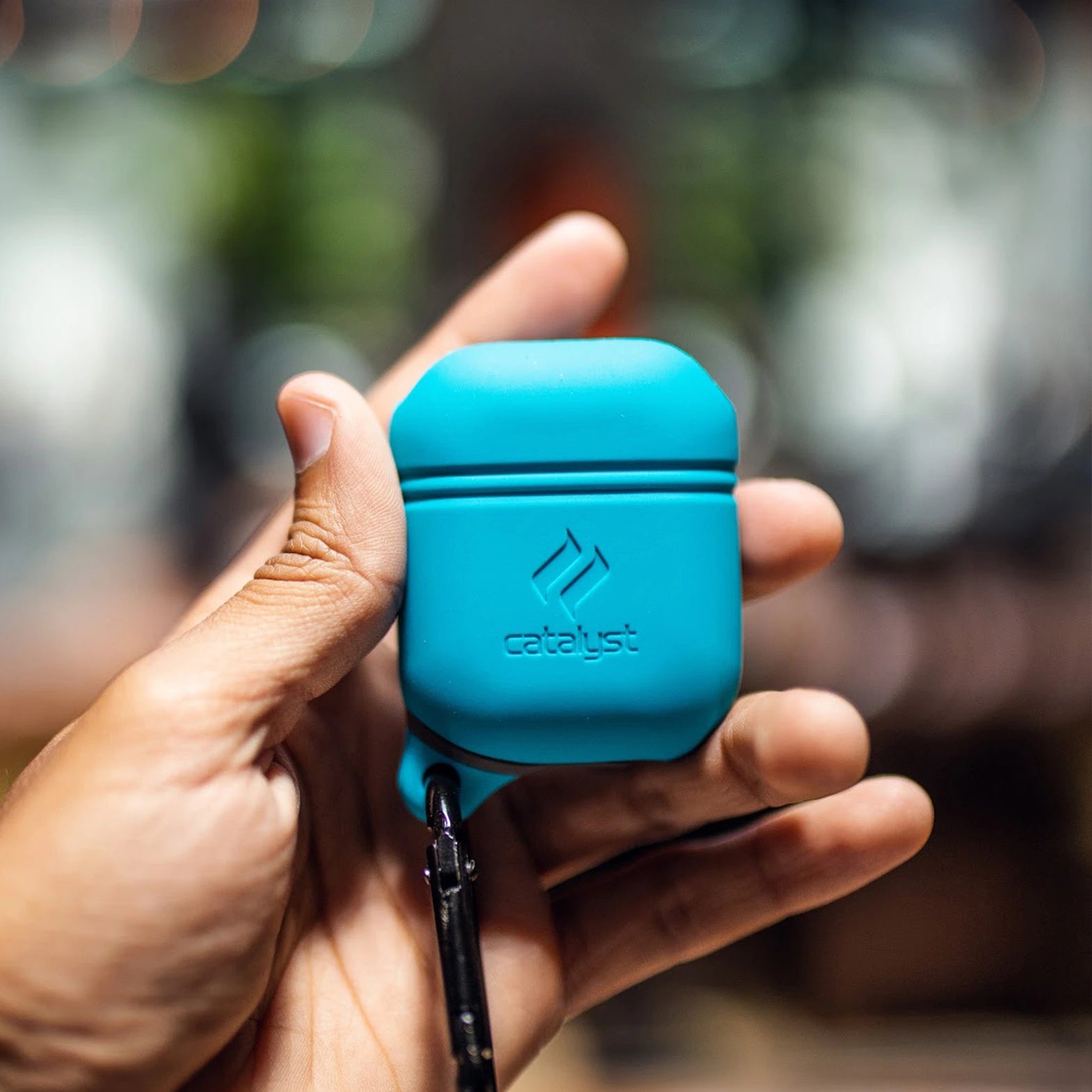 A hand holds a Catalyst waterproof case for AirPods (Gen 2/1) in blue, equipped with a small carabiner clip.