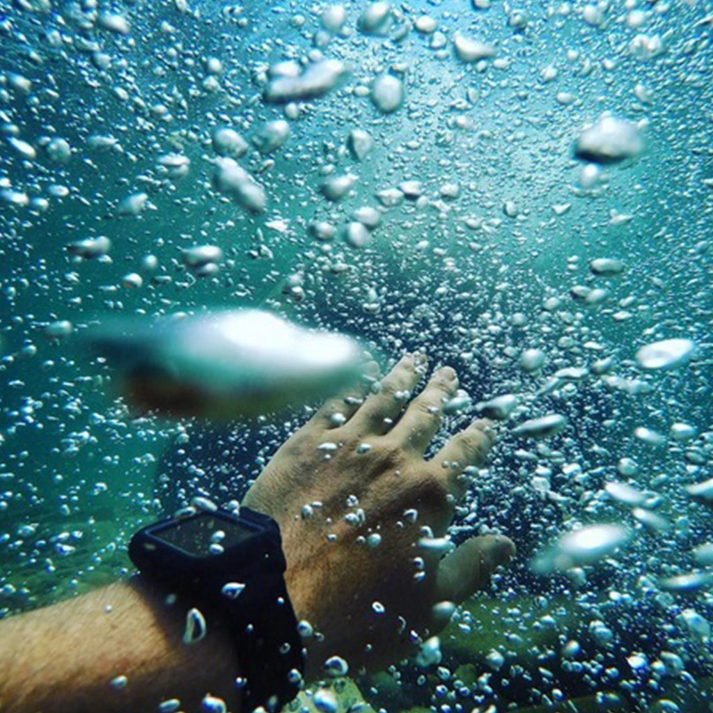 An underwater shot features a person's hand with an Apple Watch secured in a Catalyst black waterproof case, surrounded by air bubbles.