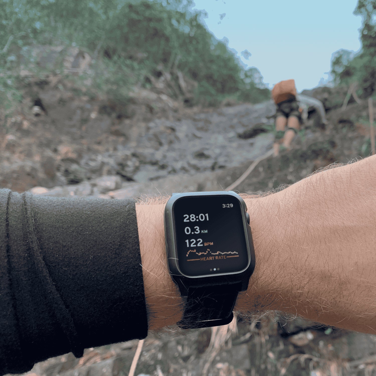 Someone with a Catalyst Apple Watch Impact Protection Case in black shows 28:01 elapsed, 0.3 km distance, and a heart rate of 122 bpm while a hiker climbs a rocky trail behind them.