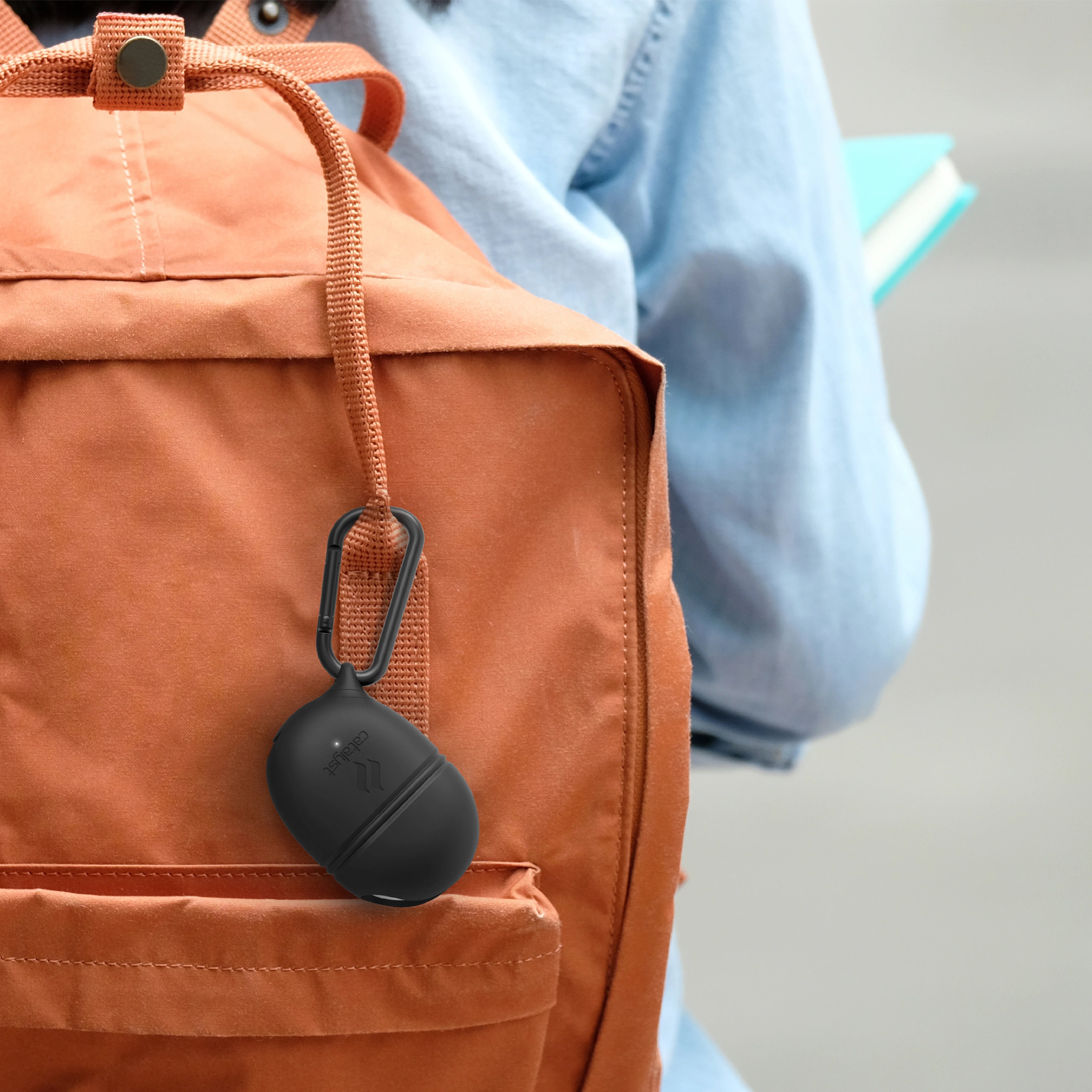 An orange backpack features a black carabiner clip holding a Catalyst Google Pixel Buds waterproof case. A person in a denim jacket carrying a teal notebook is partially visible.