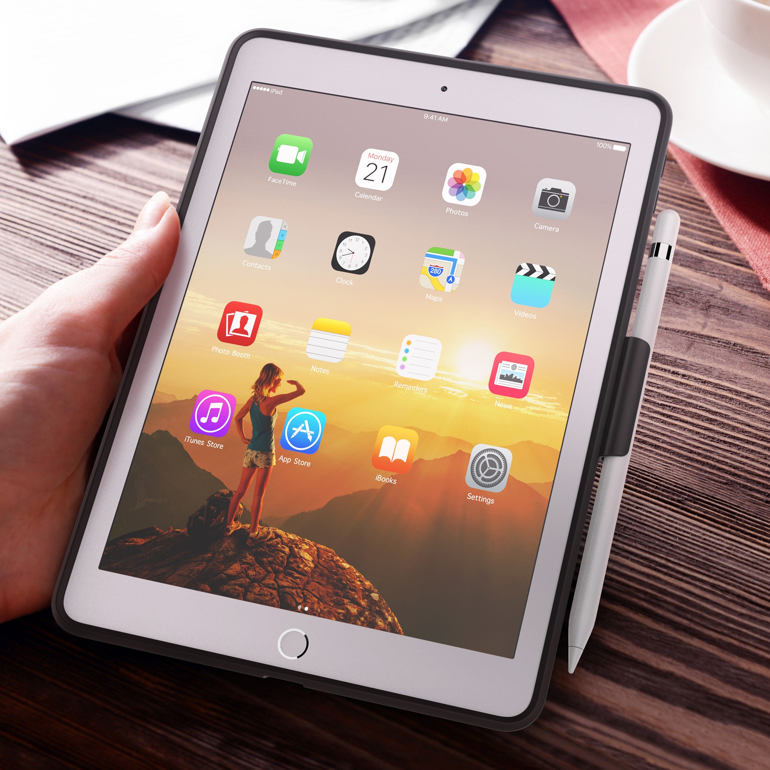 A hand holds an iPad with a sunset wallpaper and app icons, featuring a stylus attached to the side and protected by Catalyst's Impact Protection Case in black for Gen 5 & 6, ensuring it's drop proof.