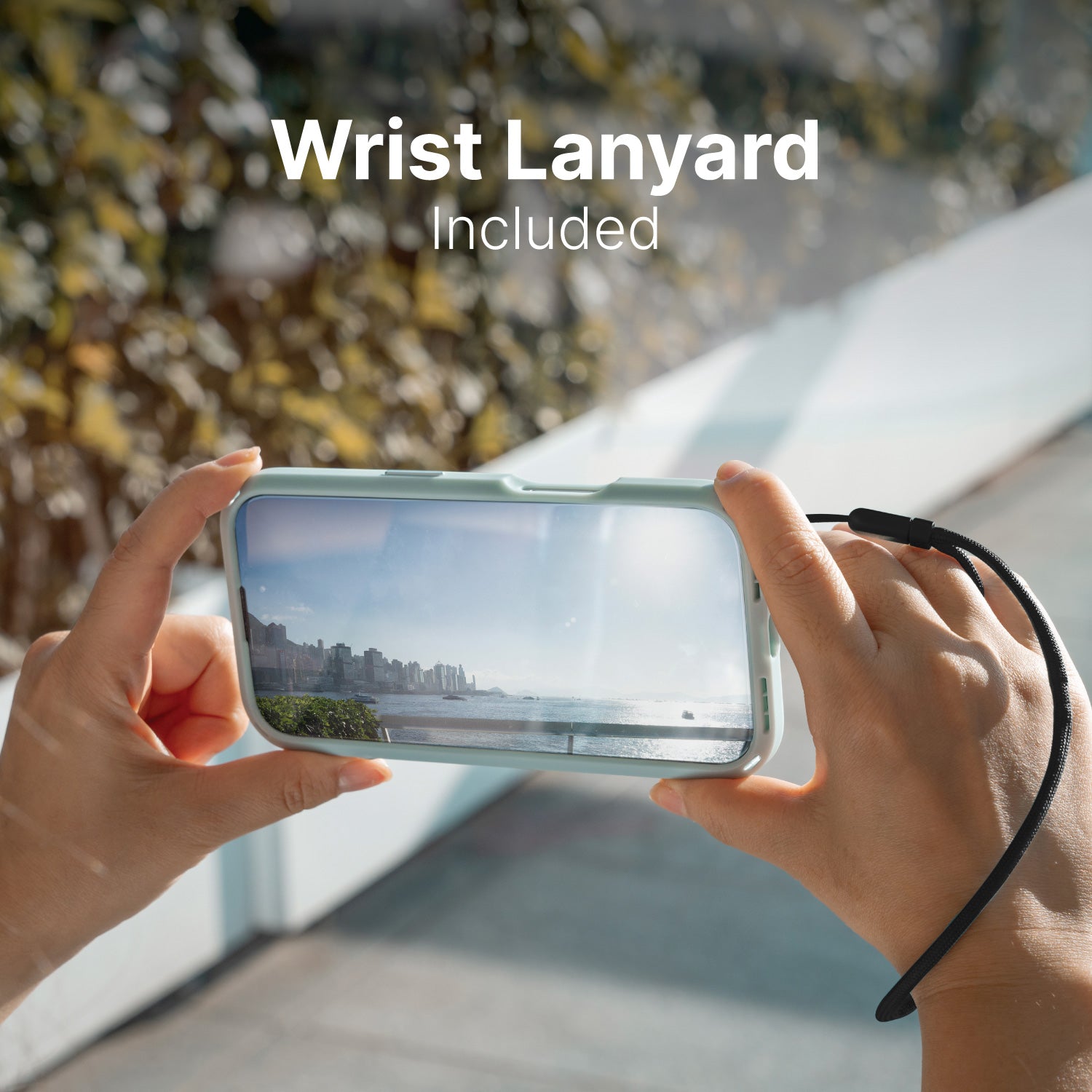 Hands holding a phone with a wrist lanyard, capturing a photo of a seaside cityscape. Text reads "Wrist Lanyard Included". The Catalyst Drop Proof Case in Sky Blue features secure lanyard attachment points for added convenience.