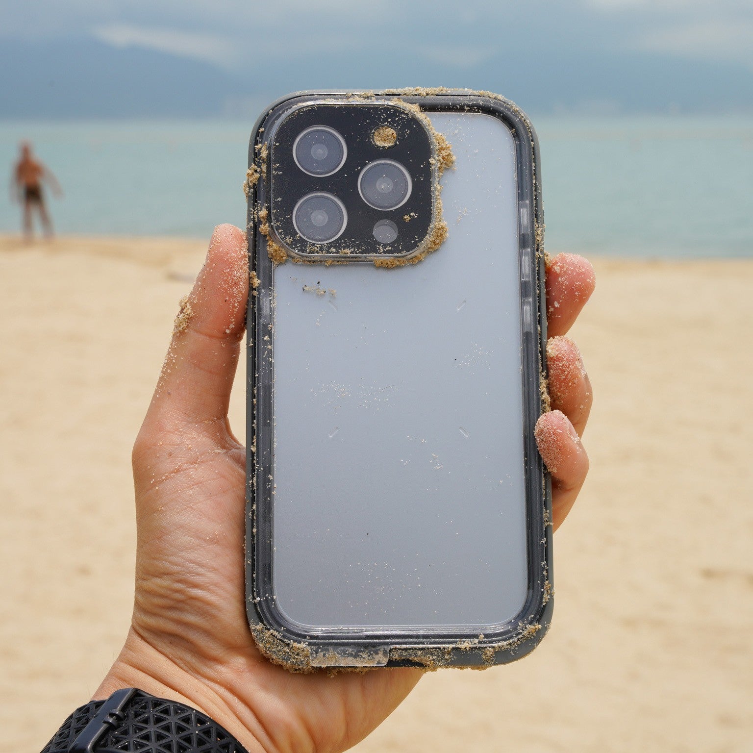 Someone grips an iPhone with a Catalyst Total Protection Waterproof Case (Black) on a beach, where waves gently lap the shore and another beachgoer meanders in the background.