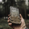 A hand holds a mud-splattered iPhone, encased in a black Catalyst total protection waterproof case, ideal for hiking adventures in the great outdoors.