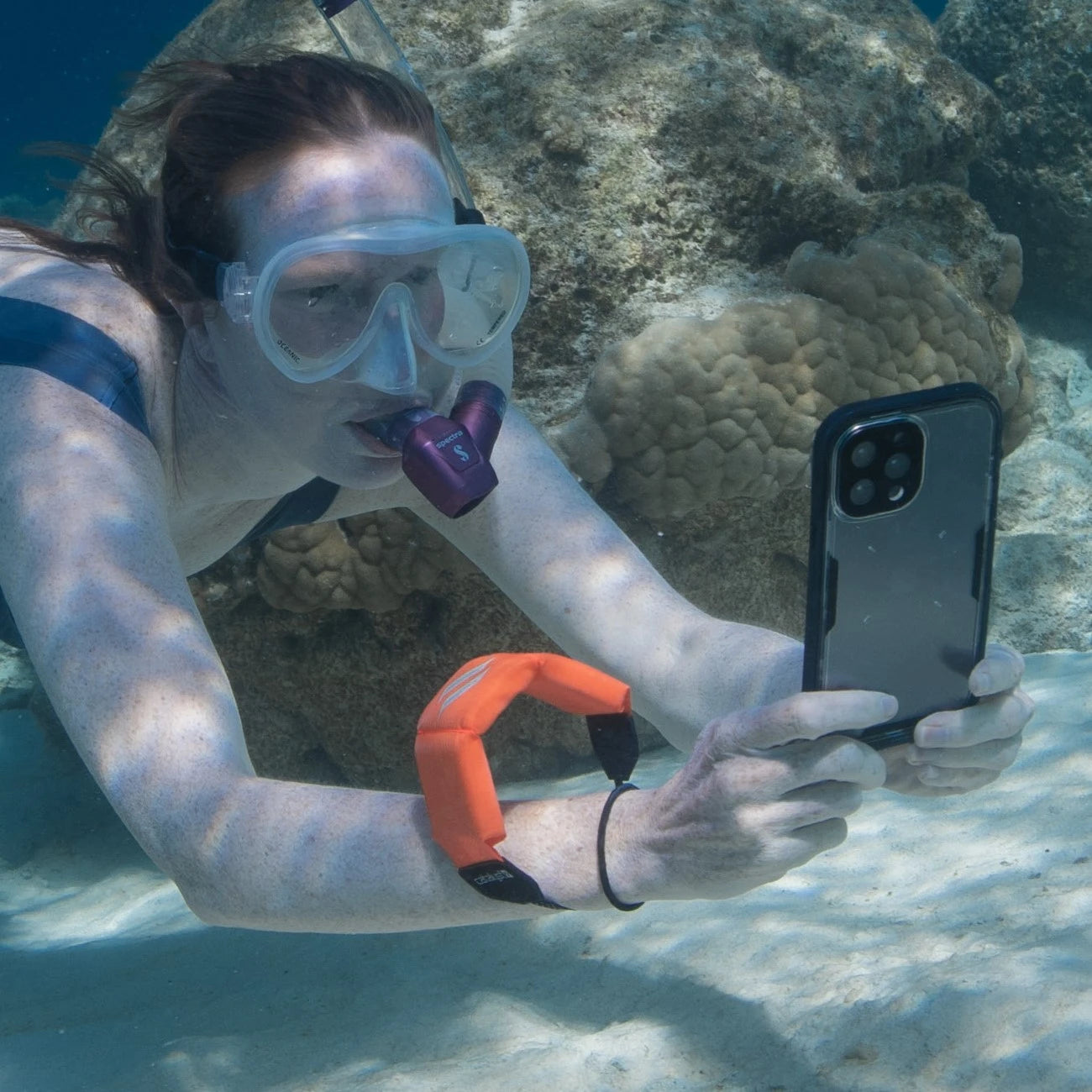 A person snorkeling underwater, equipped with goggles, a snorkel, and their iPhone secured by Catalyst's black Waterproof Case and a floating wrist lanyard. They're capturing photos of the stunning backdrop of rocks and coral.