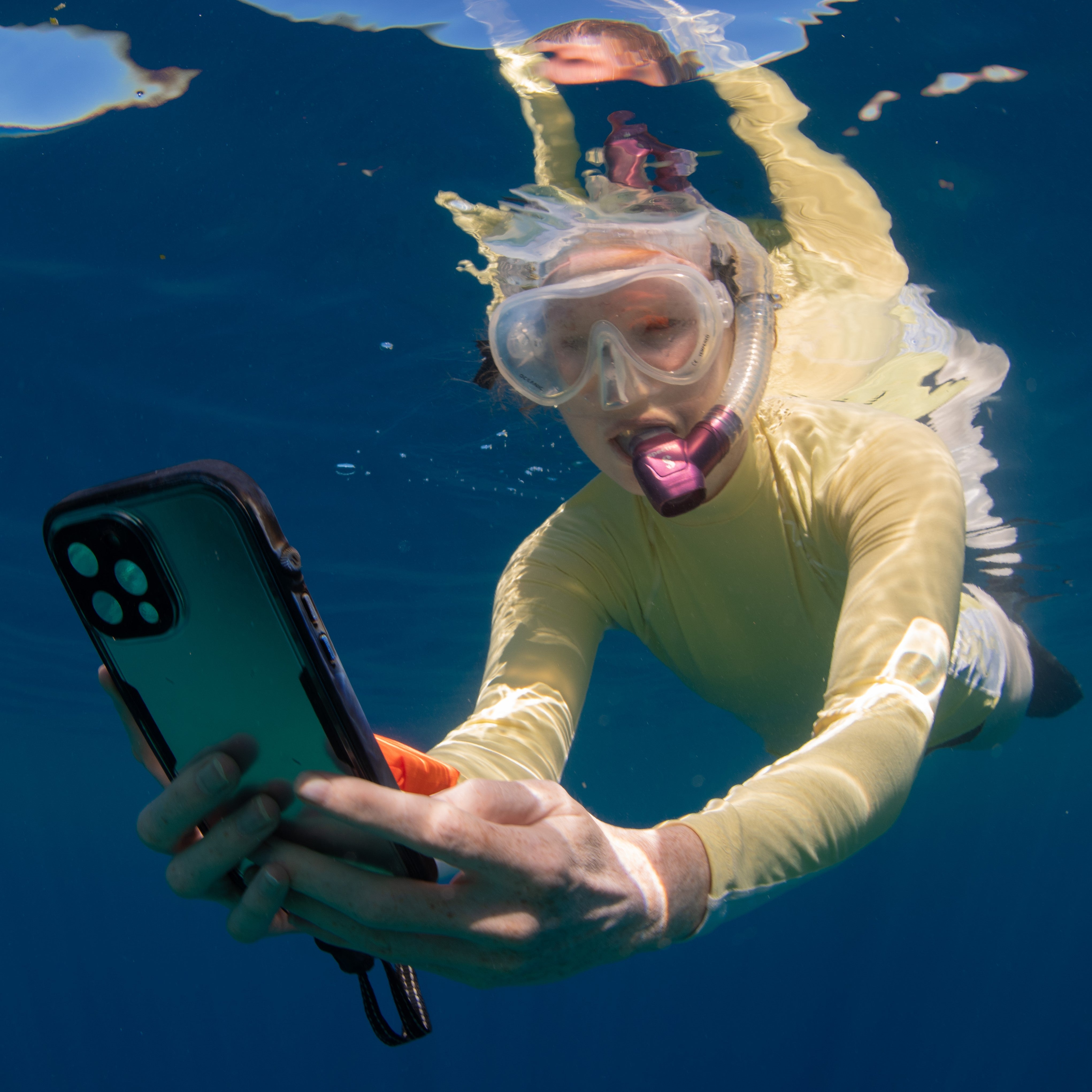 A woman snorkeling in a yellow suit and goggles captures vibrant marine life with an iPhone in a black Catalyst waterproof case.