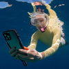 A woman snorkeling in a yellow suit and goggles captures vibrant marine life with an iPhone in a black Catalyst waterproof case.