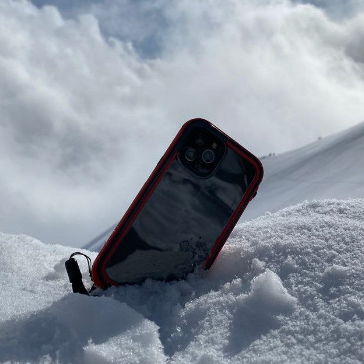 A Catalyst iPhone in a vibrant red, waterproof case offering total protection is partially buried in the snow, tilted against a snowy backdrop under a cloudy sky.