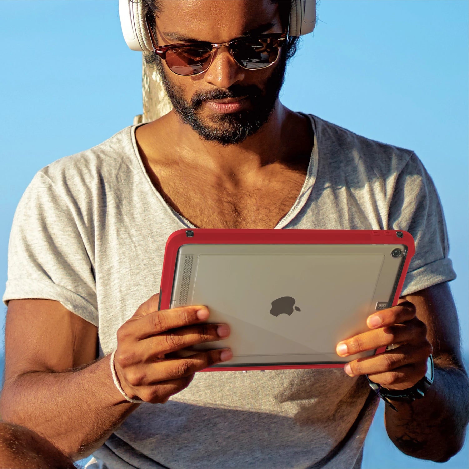 Under a clear blue sky, a person wearing sunglasses and headphones holds an iPad with a red Catalyst Waterproof Case.